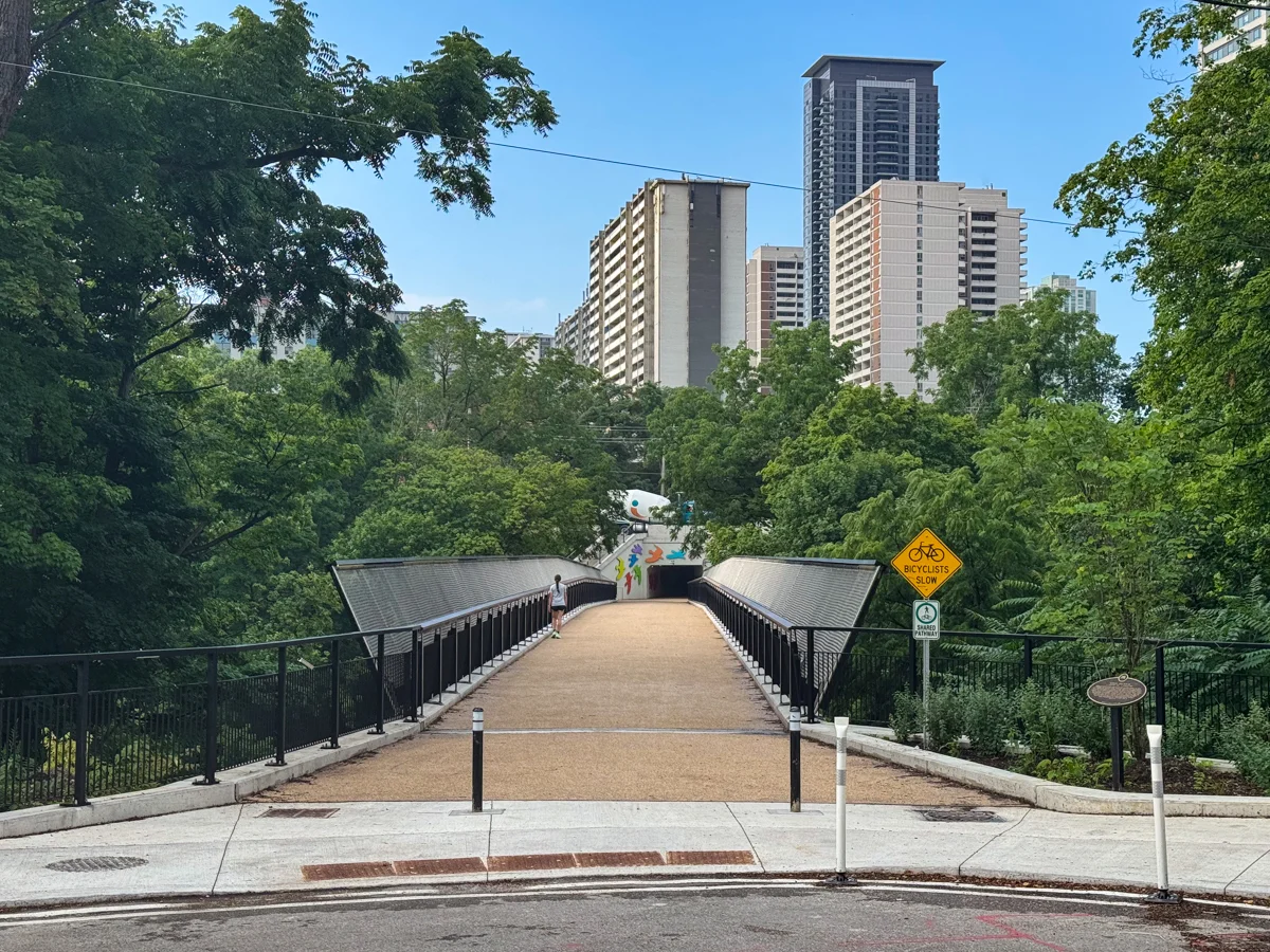 footbridge with a pedestrian, over wooded ravine with high-rises in background.