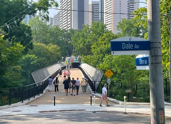 morley-callahan-bridge-2 Footbridge with multiple pedestrians over wooded ravine with high-rises in the background